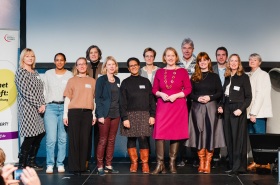 Abgebildet sind 13 Personen sie stehen auf einer Bühne vor einer weißen Wand