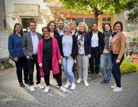 Mehrere Personen posieren für das Foto vor einem hellen gebäude. Im Hinterund ist außerdem ein Baum und Stühle zu sehen.