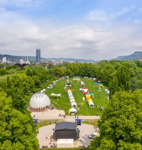 Blick auf das Fest mit Bühne, Ständen und Umweltpavillon auf der grünen Rasenmühleninsel umrandet von Bäumen, im Hintergrund der Jentower